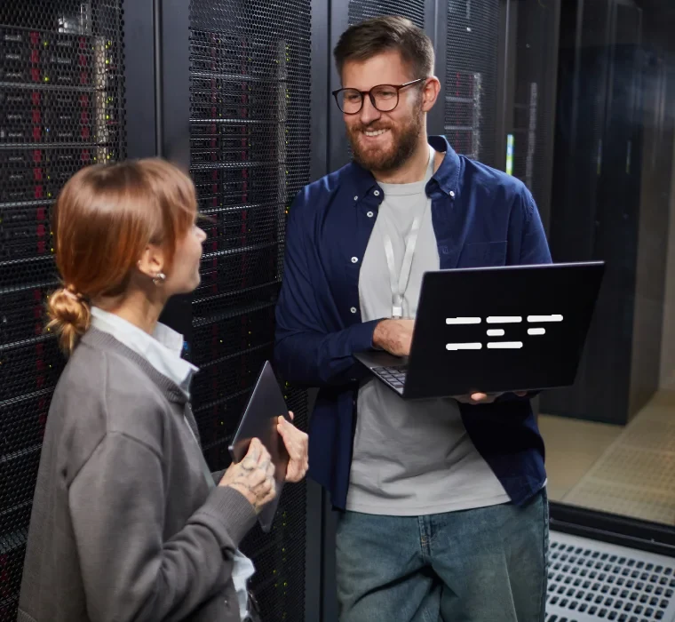 Dois engenheiros conversam ao lado de racks de servidores em um data center; um deles segura um notebook aberto.